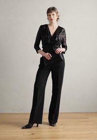 Schwarze Pailletten-Wickelbluse kombiniert mit maßgeschneiderten schwarzen Hosen. Das Model hält eine elegante schwarze Clutch und trägt spitze schwarze Pumps auf einem hellen Holzboden.