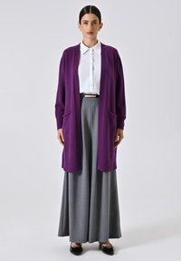 Langer lila Cardigan mit offener Front, gerippter Textur und zwei vorderen Taschen. Kombiniert mit einem weißen Hemd mit Knöpfen und grauen weiten Hosen.