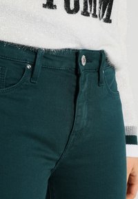 Pantalones de mezclilla en tono teal con una textura suave, que cuentan con un cierre frontal de botón, lazos para cinturón y bolsillos laterales. Acentuados por un suéter blanco suave.