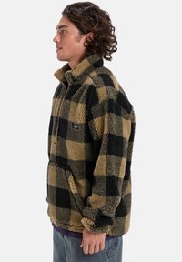 Fleece pullover in a black and tan checkered pattern. Features a high collar, quarter-zip, and loose fit with elasticated cuffs.