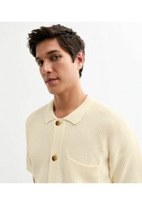 Gebreide polo shirt in lichtbeige met een gestructureerde afwerking, korte mouwen, een kraag en een enkele borstzak. Voorzien van een knoopsluiting.