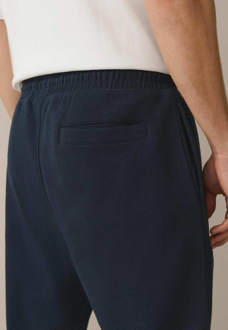 Uomo che indossa pantaloni blu navy con vita elasticizzata e tasca posteriore a filetto, e una camicia bianca, primo piano sulla zona della vita e dei fianchi.