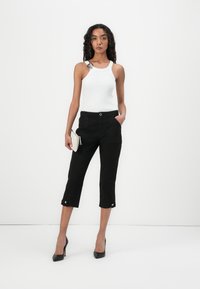 Top sin mangas blanco con canalé y tirantes ajustables, combinado con pantalones cropped negros que tienen bolsillos frontales y dobladillo abotonado, usados con tacones negros.