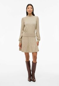 Maglione beige a maglia con collo alto e maniche a sbuffo, abbinato a una gonna svasata dello stesso materiale. Indossato con stivali marroni fino al ginocchio.