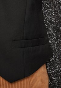 Černý blazer s texturovaným povrchem, vybavený kapsou na švu a špičatým klopem, v kombinaci s třpytivou šedou tkaninou a okrově hnědým velurem.
