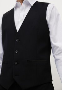 Gilet nero su misura realizzato in tessuto texturizzato, dotato di cinque bottoni, tasche laterali e un design nitido e strutturato. Indossato sopra una camicia bianca.