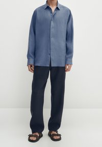 Ljusblå skjorta med långa ärmar, kombinerad med mörknavy lösa byxor och bruna sandaler. Slät tygtextur.