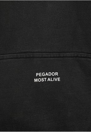 Melns audums ar baltu izšūtu tekstu "PEGADOR MOST ALIVE" virs horizontālas šuves līnijas.