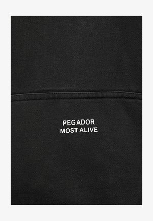 Fekete anyag fehér hímzett felirattal: "PEGADOR MOST ALIVE" egy vízszintes varrásvonal fölött.