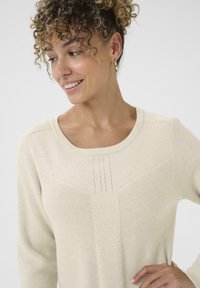 Világos bézs pulóver texturált mintával, széles nyakkivágással és finom függőleges vonalakkal. Az anyag puhanak és kötöttnek tűnik.