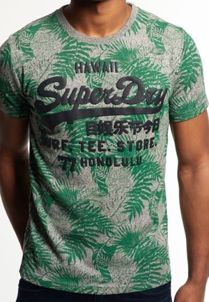 Man wearing gray and green leaf-patterned T-shirt with black text "SuperDry," "Hawaii," and "Honolulu" on the front.