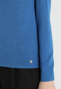 Blå långärmad tröja i mjuk väv med en silverfärgad logotyp på nederkanten och en figurnära design. Svart plagg under.