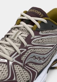 Sneaker avec une tige en mesh beige, accents bordeaux, détails argentés, lacets blancs épais et une semelle contrastante noire et jaune.
