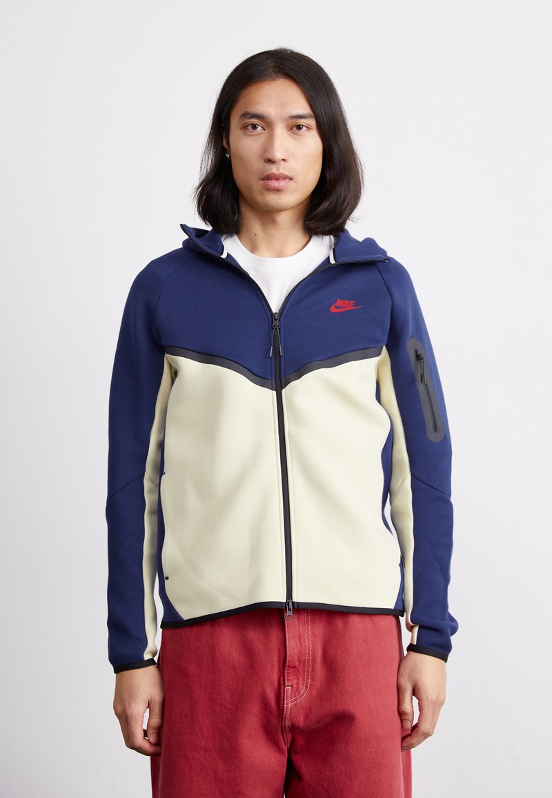 Nike zip-up hættetrøje i navy og creme, med et kontrastdesign, frontlynlås, slim fit og sidelomme med lynlåslukning.