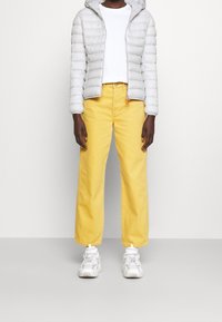 Doudoune gris clair avec capuche, t-shirt blanc à col rond, et pantalon jaune vif à jambes larges. Baskets blanches avec des accents texturés.