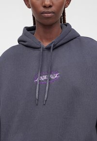 Gros plan d’une personne portant un sweat à capuche gris foncé avec des cordons tressés et un petit motif brodé violet et blanc sur la poitrine.