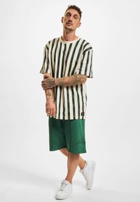 Chemise à manches courtes rayée couleur crème avec des lignes verticales vertes, associée à un short vert. Complétée par des baskets blanches et des tatouages visibles.