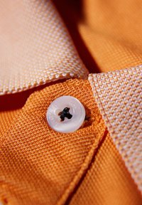 Pomarańczowa teksturowana tkanina z białym guzikiem z czarną nitką. Zbliżenie na krawędź kołnierzyka, podkreślające detale materiału i szycia.