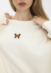 Felpa bianca con polsini a coste, con una farfalla ricamata arancione e nera sul davanti. Tessuto morbido, design casual.