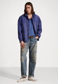 Μπλε βαμβακερό μπουφάν με κουμπιά και τσέπη, φορεμένο πάνω από ριγέ αμάνικη μπλούζα. Φθαρμένο ανοιχτό μπλε τζιν και λευκά αθλητικά ολοκληρώνουν την εμφάνιση.