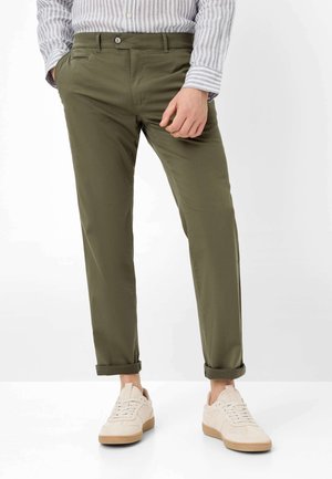 Homme portant un pantalon olive retroussé, une chemise à rayures claire boutonnée et des baskets beige, debout avec une main dans la poche.