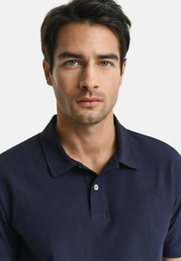 Polo-shirt bleu marine en tissu texturé, doté d'une patte de boutonnage à deux boutons et d'une broderie de logo subtile sur la poitrine. Manches courtes.