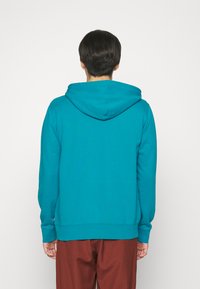 Turquoise pullover hoodie met een trekkoordkap, lange mouwen en ribgebreide manchetten. De stof lijkt glad. Gemaakt met een paar bruine broeken.