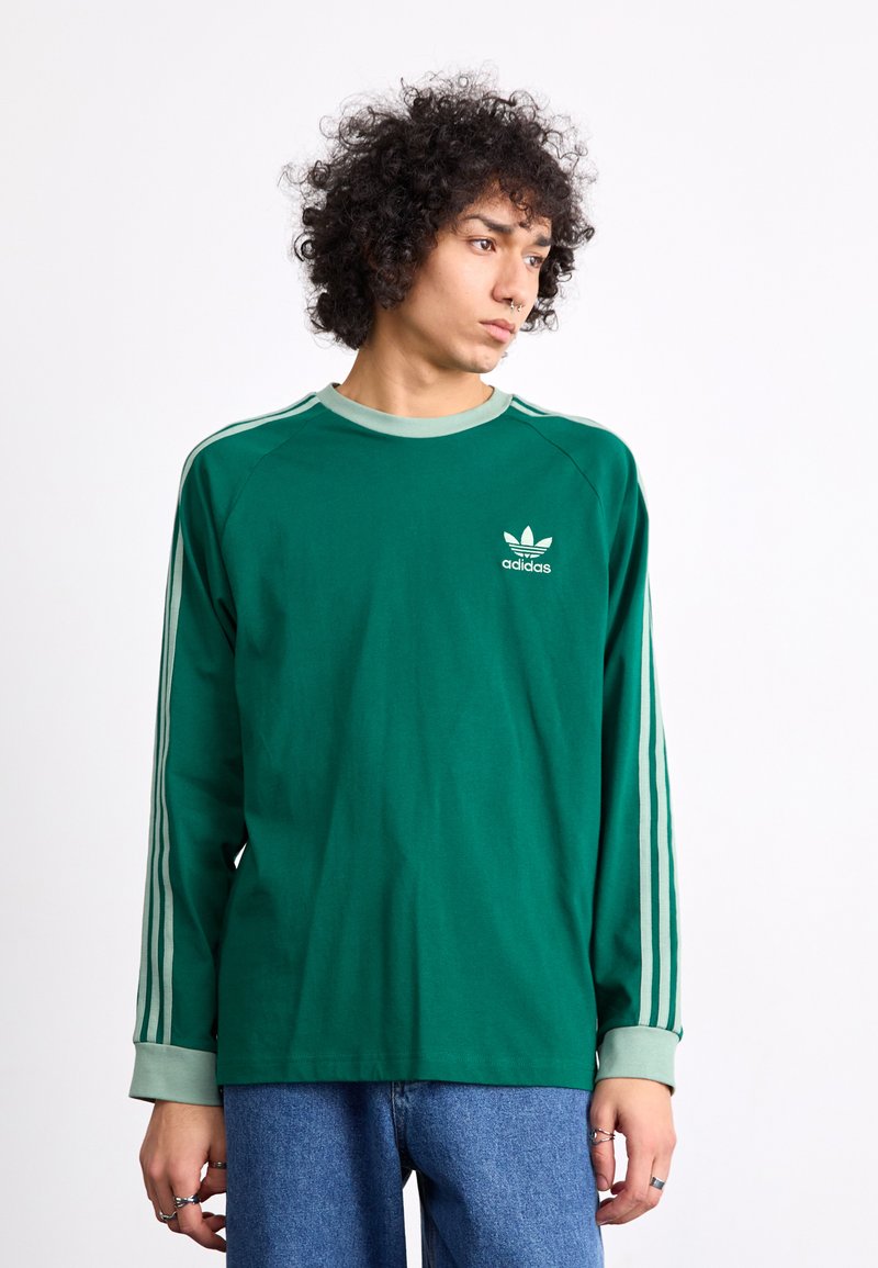 Ilga žalia marškinėlių su ilgomis rankovėmis su šviesiai žaliai akcentais, su mažu baltu Adidas logotipu ant krūtinės ir trimis baltais dryžiais ant rankovių.
