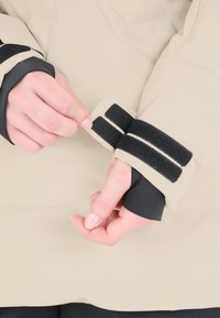 Beige vattentät jacka med manschett som har svarta kardborreband och en slät yta. Händer justerar manschetten med en synlig svart inre foder.