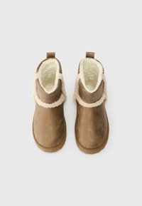 Botas castanhas até ao tornozelo com um material exterior suave, forro bege felpudo e uma tira decorativa trançada à volta do tornozelo. Bico redondo, sola texturizada.