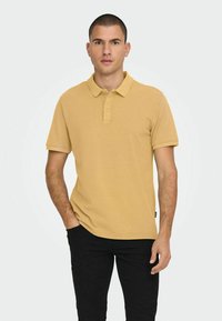 Polo de color mostaza hecho de algodón, con cuello, tapeta de dos botones y mangas cortas, combinado con pantalones negros.