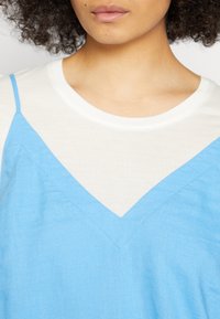 Top sem mangas azul claro sobre uma camisa branca, apresentando um decote em V e um tecido texturizado, com costuras visíveis e linhas limpas.