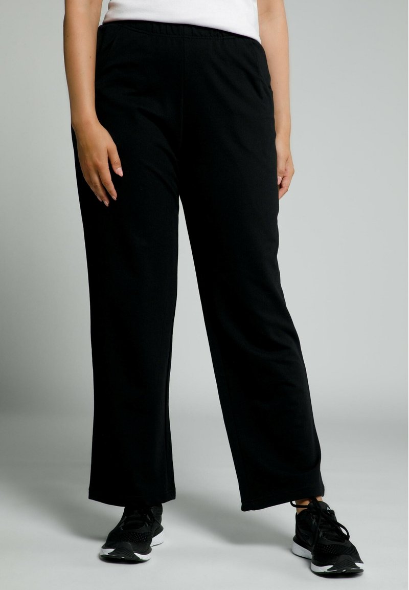 Pantalones de chándal negros con cintura elástica, de diseño suelto y pierna recta. La tela parece suave y cómoda, combinada con zapatillas deportivas negras.