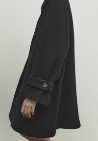 Sort, oversized frakke i tekstureret uldstof. Har brede ærmer og knapdetaljer på manchetterne. Enkel, struktureret silhuet.