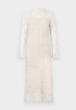 Biała koronkowa sukienka z długim rękawem o falistym wzorze, z wysokim dekoltem i półprzezroczystą tkaniną na podszewce, prezentowana na jednolitym tle.