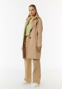 Beige wollen jas met een dubbele rij knopen, grote knopen, klepzakken en een capuchon, gedragen over een groene top en beige broek. Witte sneakers.