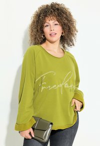 Sweat à capuche vert à manches longues, avec "Liberté" brodé en blanc. Assorti à une pochette grise, porté avec un jean foncé.