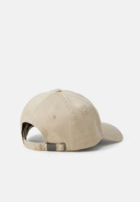 Beige caps laget av bomull med buet skygge, justerbar stropp og metallspenne. Har en sydd knapp og ventilasjonsøyeler.