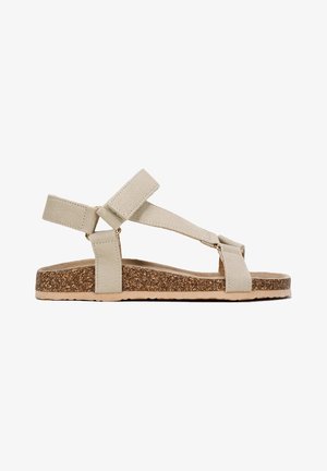 Beige suede sandals with a cork sole, featuring two adjustable straps and a flat design for comfort. Notable gold hardware accents included.