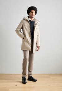 Parka beige con cierre de cremallera, dos bolsillos en el pecho y capucha. Lleva puesto un suéter oscuro y combina con pantalones beige y zapatos negros.