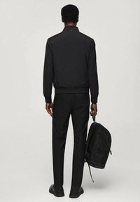 Zwarte bomberjack met een hoge kraag, gecombineerd met slim fit zwarte trousers. Persoon houdt een zwarte rugzak vast en staat tegen een neutrale achtergrond.