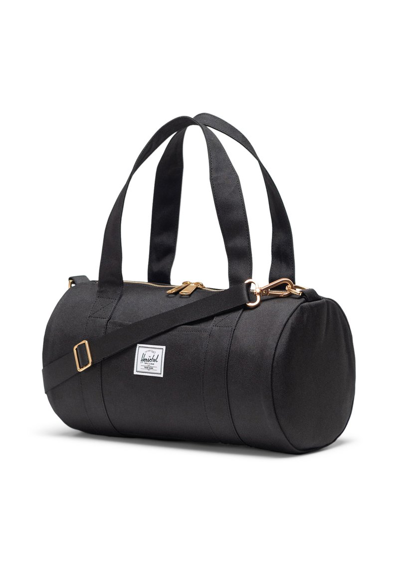 Crni cilindrični putni kofer s dvostrukim ručkama na vrhu, podesivim ramennim remenom, zlatnim patentnim zatvaračem i bijelom Herschel oznakom s strane.