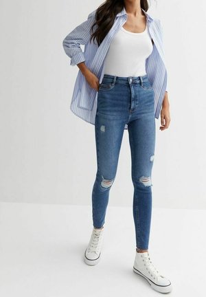 Mujer con vaqueros ajustados rasgados de color azul, camiseta sin mangas blanca, camisa de botones a rayas azul claro y zapatillas altas blancas.