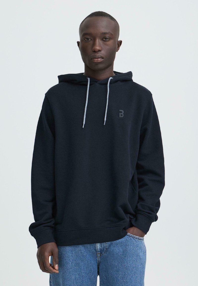 Navy hoodie met trekkoorden, logo op de borst en geribbelde boorden, gecombineerd met lichtblauwe jeans. Zachte stof en casual pasvorm.