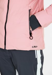 Rosa puffjacka med svart dragkedja och CMP-logotyp. Har sidofickor och elastiska muddar, tillsammans med mörka byxor som framhäver vita ränder.