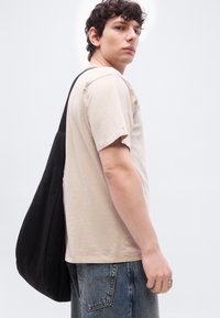 Νεαρός άνδρας που φοράει μπεζ μπλουζάκι και μπλε τζιν, κρατώντας μια μεγάλη μαύρη τσάντα ώμου, κοιτάζει πάνω από τον ώμο του με φόντο απλό.