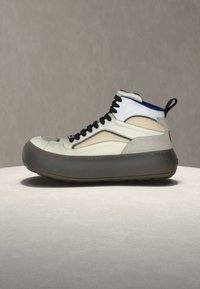Vysoký teniskový topánok s béžovými a bielymi panelmi, čiernymi šnúrkami, hrubou čiernou podrážkou a modrou podšívkou, vystavený na svetlom povrchu proti sivému pozadiu.