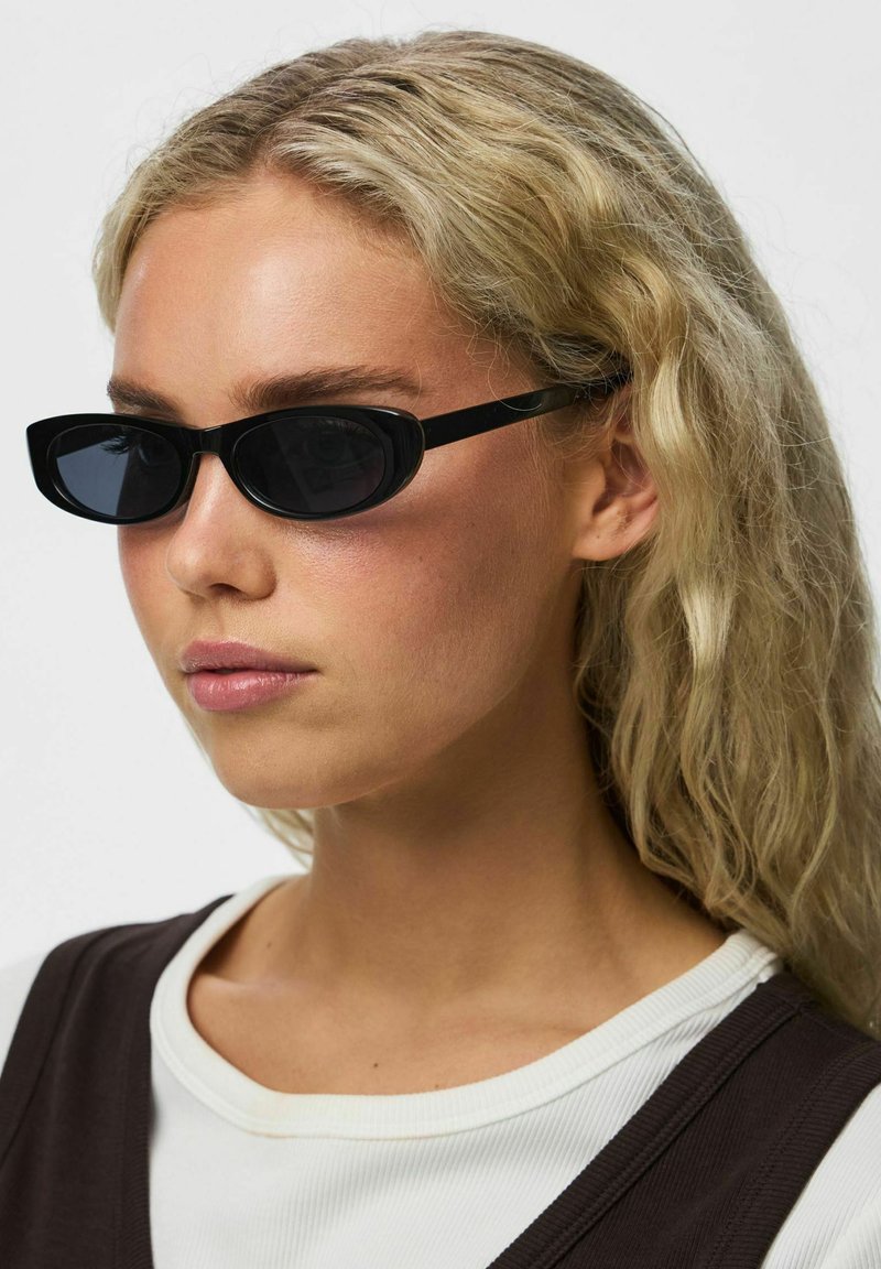 Femme aux cheveux blonds ondulés portant des lunettes de soleil noires fines et une chemise blanche à plusieurs couches avec un top sans manches foncé, légèrement tournée vers la gauche.
