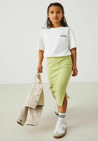 Witte T-shirt met korte mouwen met de opdruk "LEMONADE", geribde groene midi rok, lichtbruine denim jack, witte en grijze sneakers met zwarte accenten.
