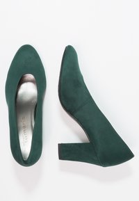 Gröna mocka högklackade skor med runt tåparti, med ett urtaget mönster och en silverfärgad innersula. Klack i blockstil ger stabilitet.